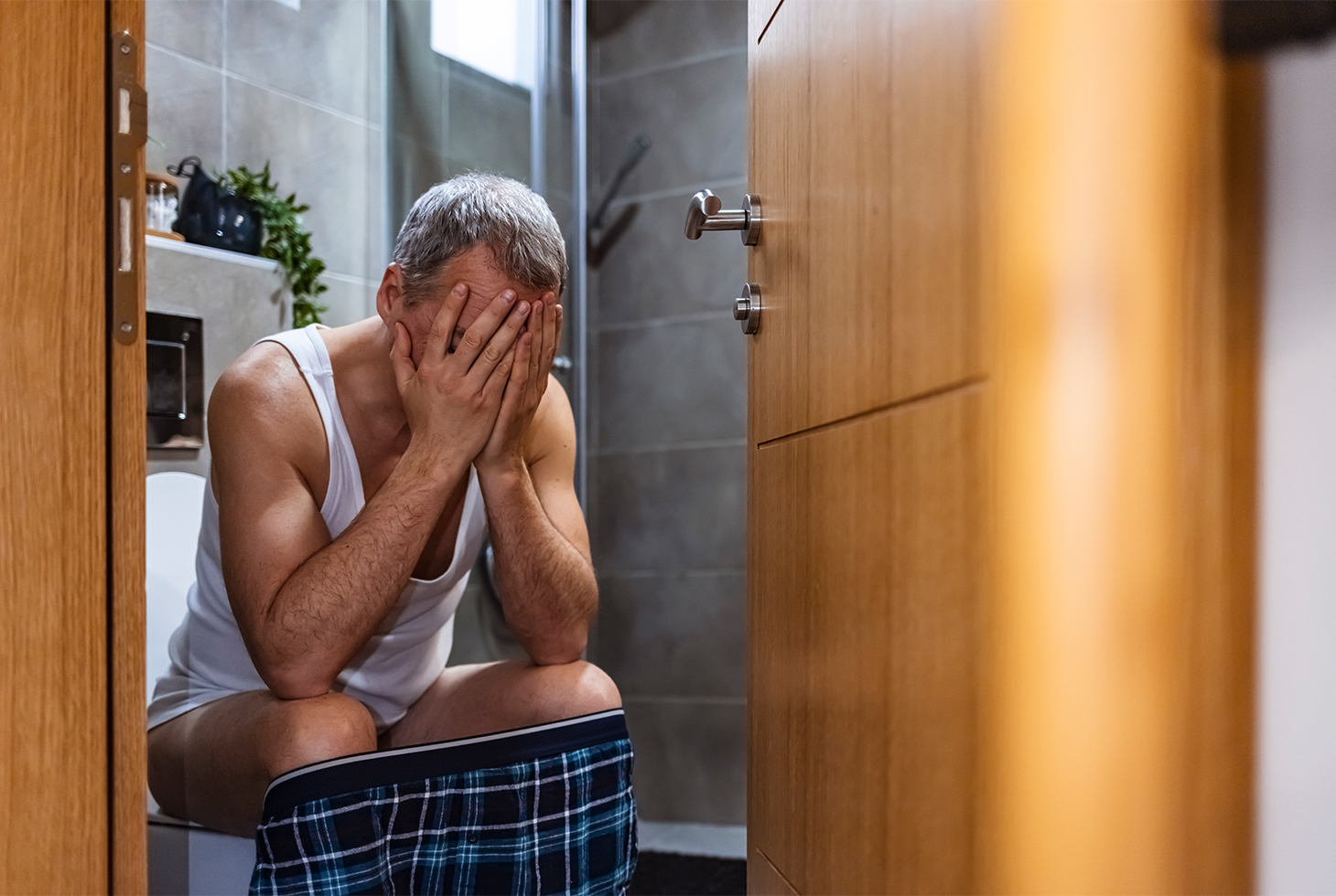 Un homme senior assis sur les toilettes se tenant la tête entre les mains. Visuel pour illustrer la constipation chronique et ses risques liés. 