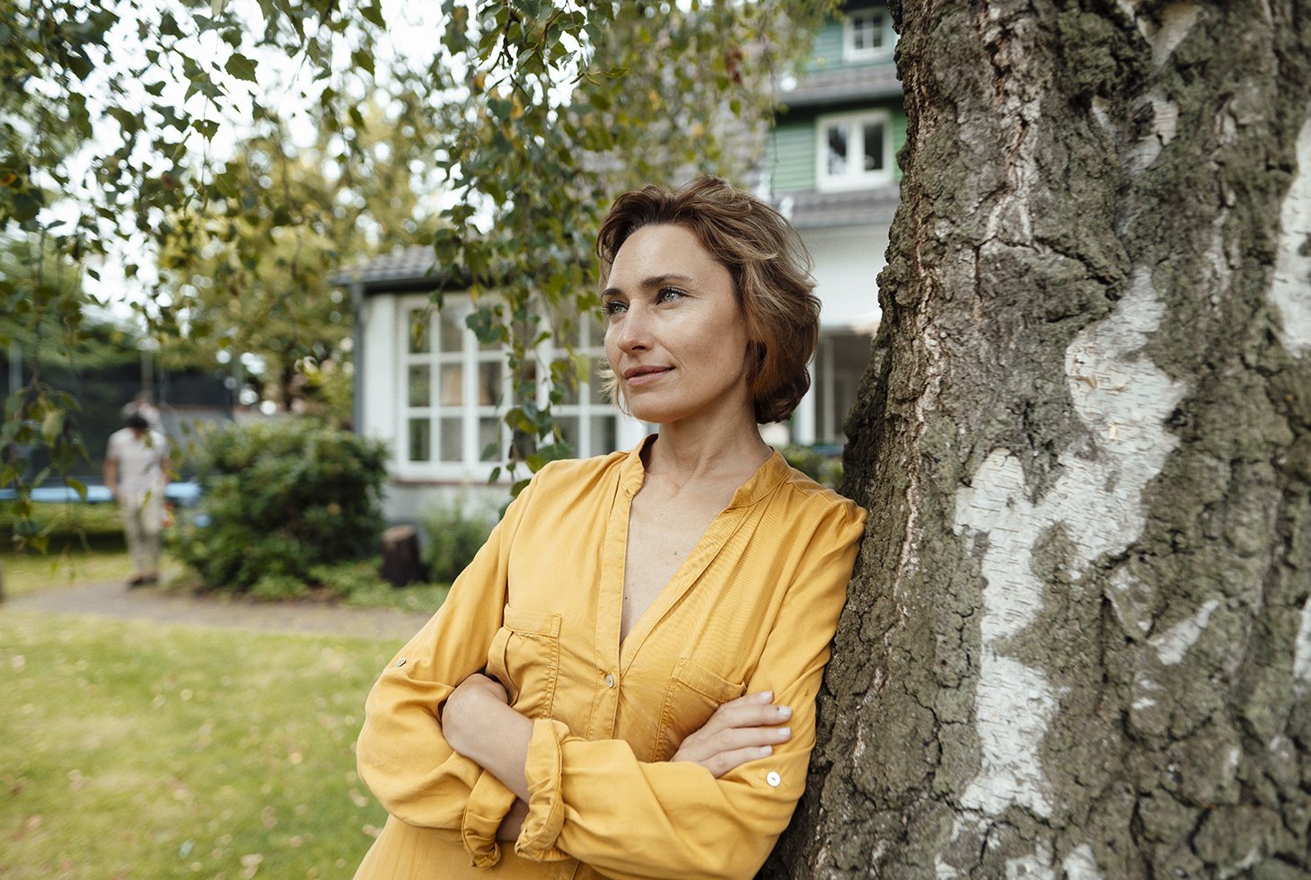Portrait d’une femme les bras croisés, adossée à un tronc arbre devant une maison. Image pour illustrer la thématique incontinence mixte.