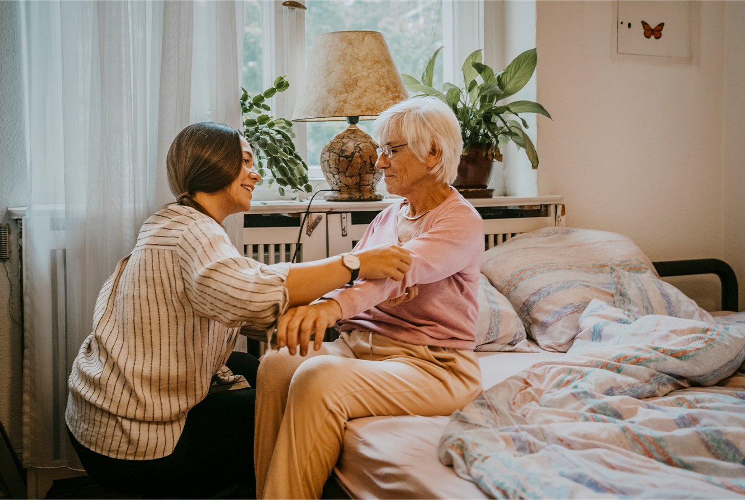 Une personne âgée est assise sur son lit, une aidante l’aide à s’habiller. Visuel pour illustrer l'accompagnement de personnes dépendantes en situation d’incontinence.