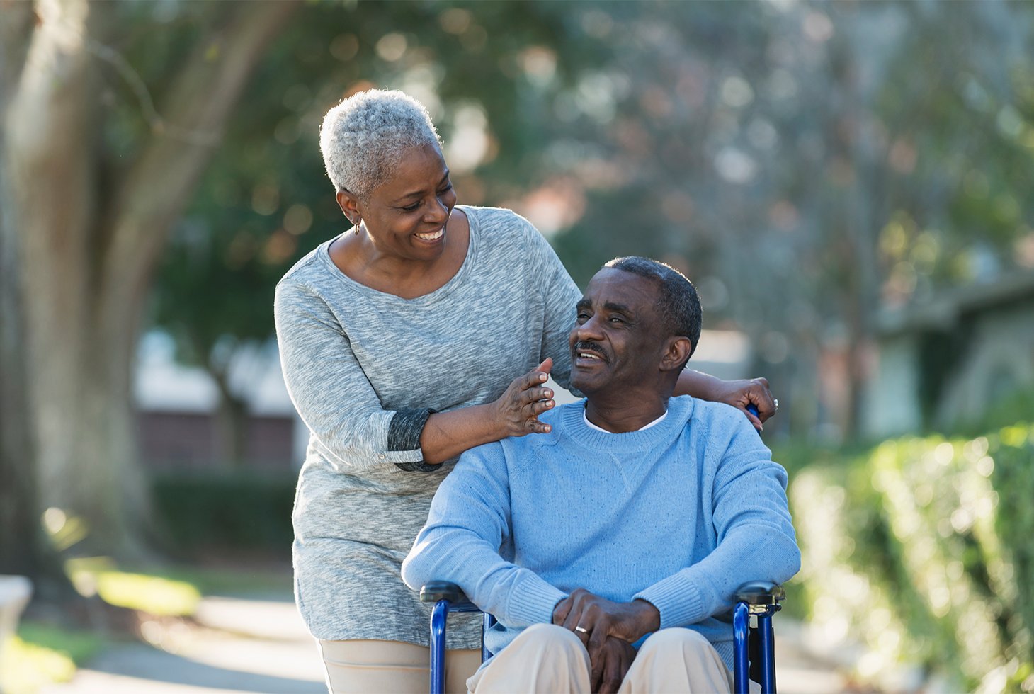 Portrait d'un couple senior lors d’une promenade. L'homme est assis dans un fauteuil roulant poussé par sa femme. Ils parlent et sourient.