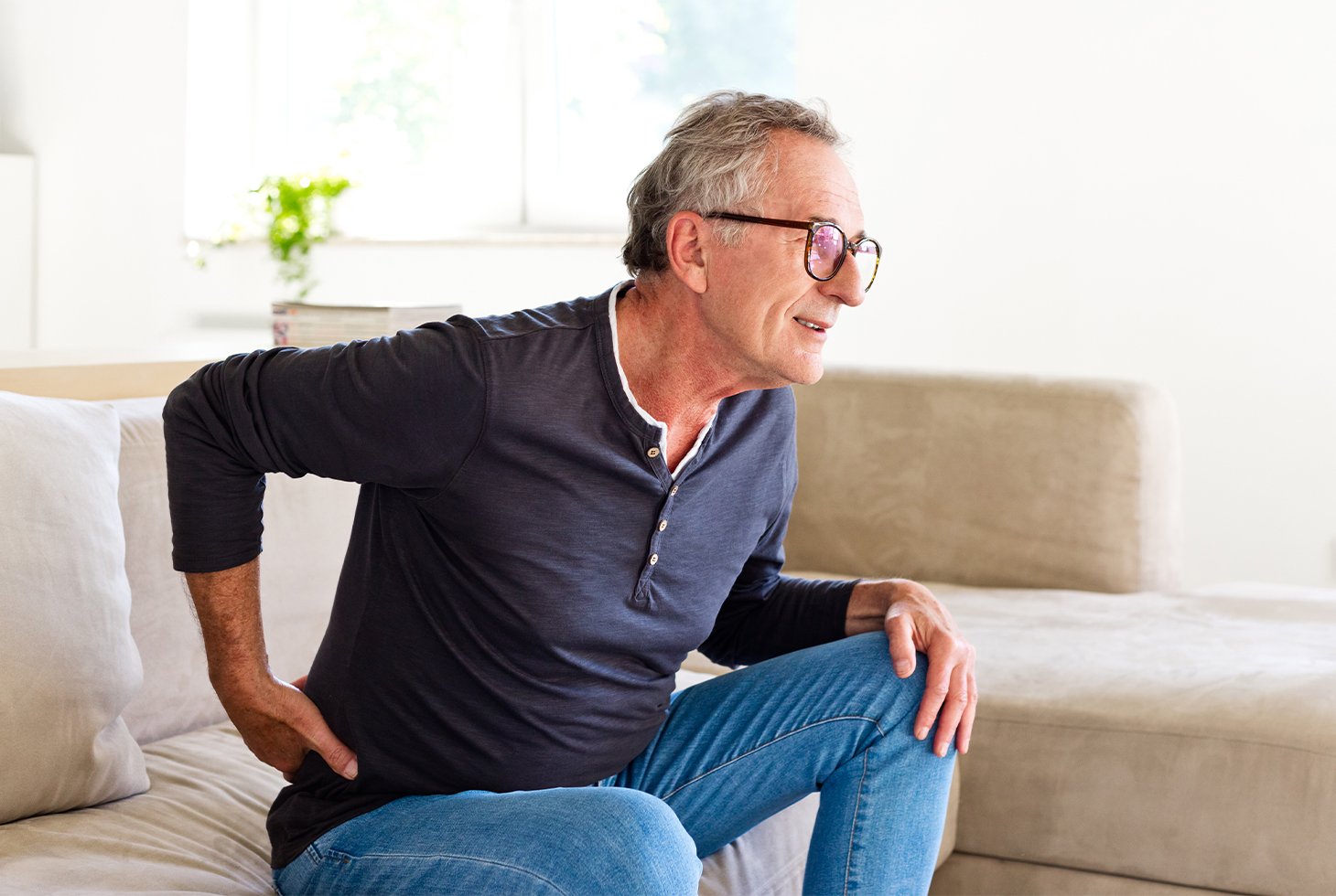 Un homme, assis sur un canapé, se tient le bas du dos. Visuel pour illustrer le risque d’incontinence urinaire liée à une affection chronique.