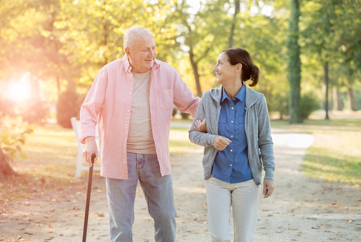 Portrait d’une personne âgée et son aidante lors d’une promenade. L’homme marche à l’aide d’une canne et tient par le bras sa proche-aidante