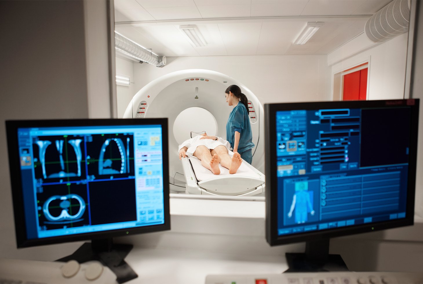 Photo d’un examen d’imagerie médicale : un homme est en position allongée sur un lit d’examen avec debout, à sa droite, une manipulatrice en électroradiologie médicale.